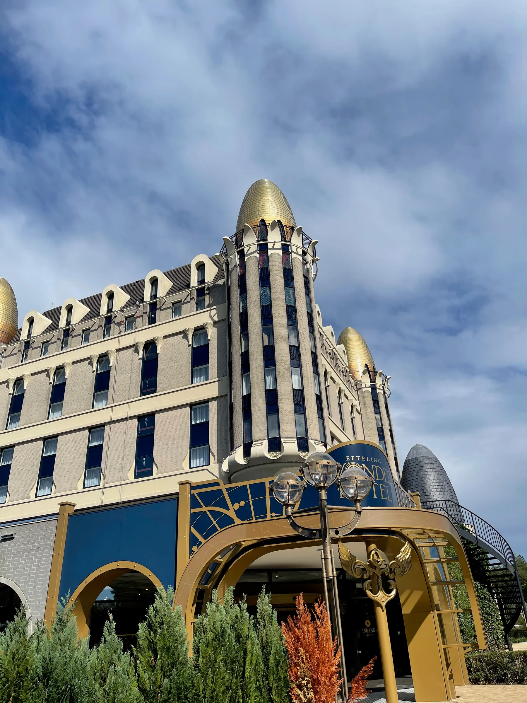 Prédio em estilo arquitetônico com torre dourada pontiaguda, fachada bege, detalhes em azul e dourado, céu parcialmente nublado e arbustos na frente.