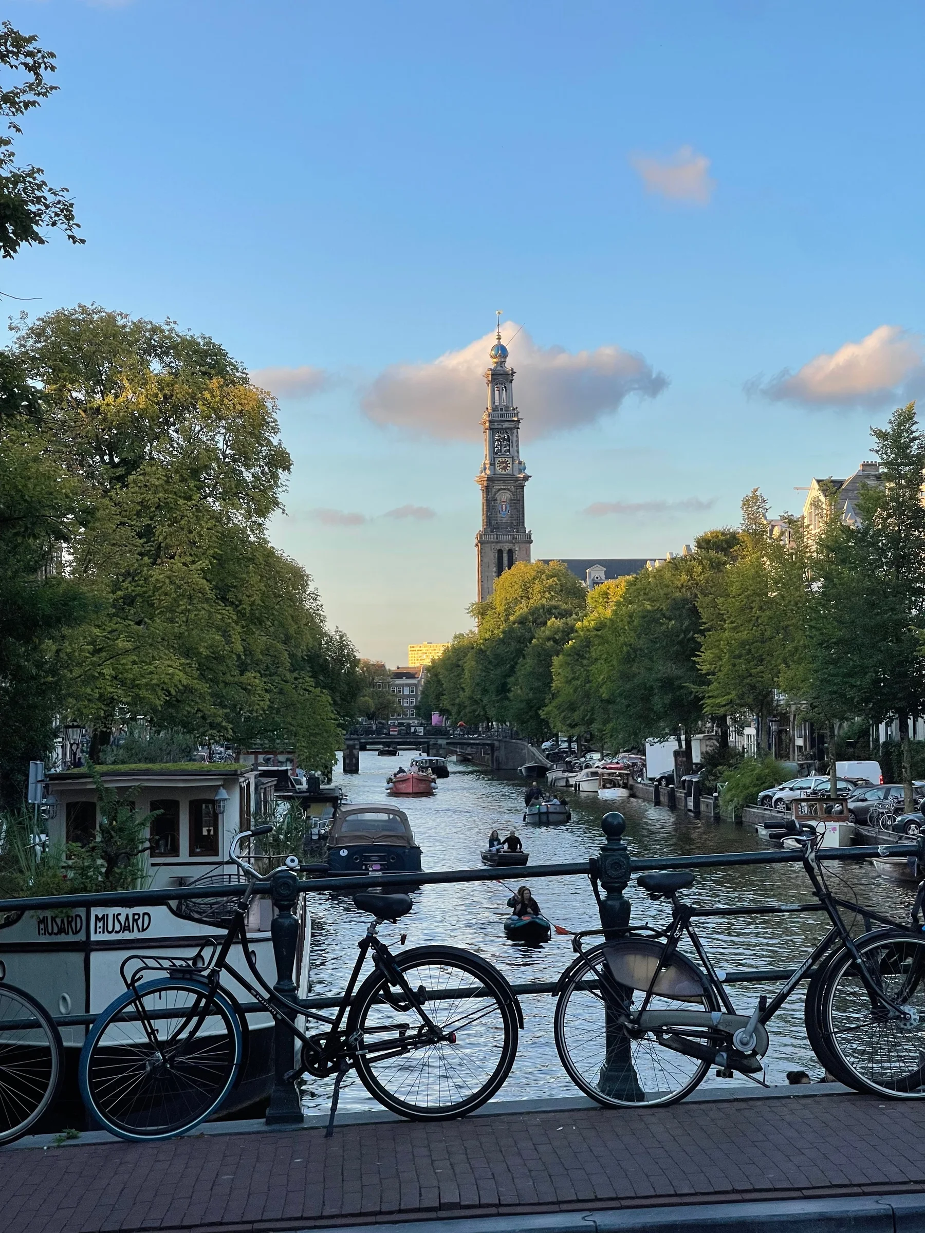 Três bicicletas encostadas em grade de ponte sobre canal, com barcos e árvores ao redor e torre de igreja ao fundo sob céu azul com nuvens.