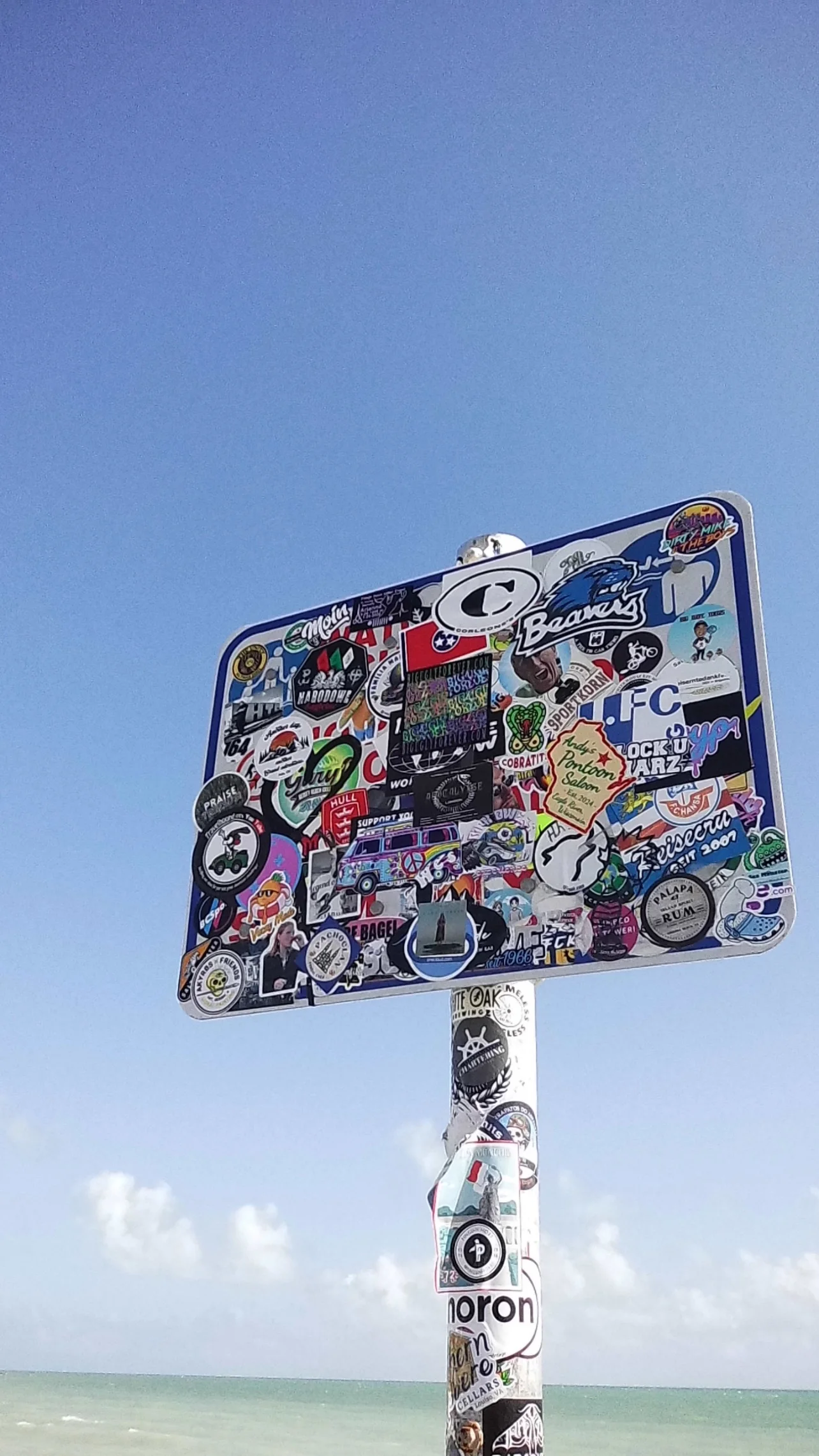 Placa metálica retangular repleta de adesivos coloridos fixada em poste, com céu limpo e mar ao fundo