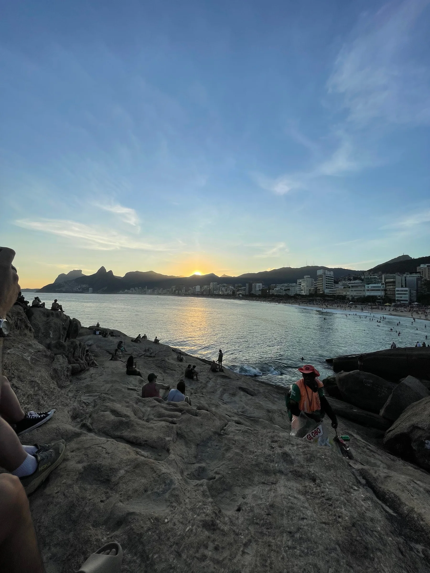 Pessoas sentadas em pedras à beira-mar contemplam o sol se pondo atrás de montanhas e edifícios ao longe, sob céu claro.