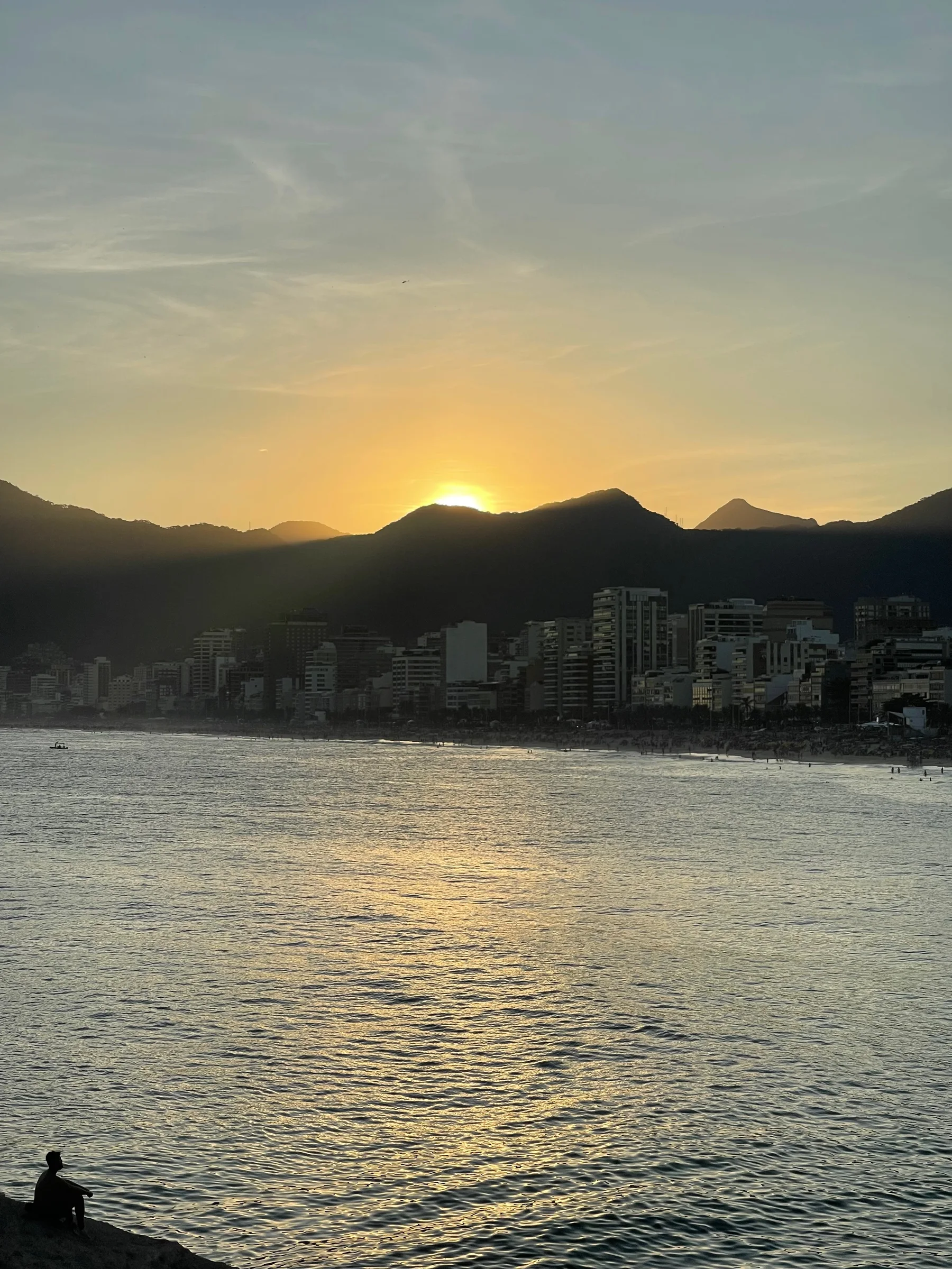 Pôr do sol ao fundo de montanhas com sol parcialmente oculto, mar com reflexo dourado, prédios urbanos silhuetados e uma pessoa sentada em pedra próxima à água.